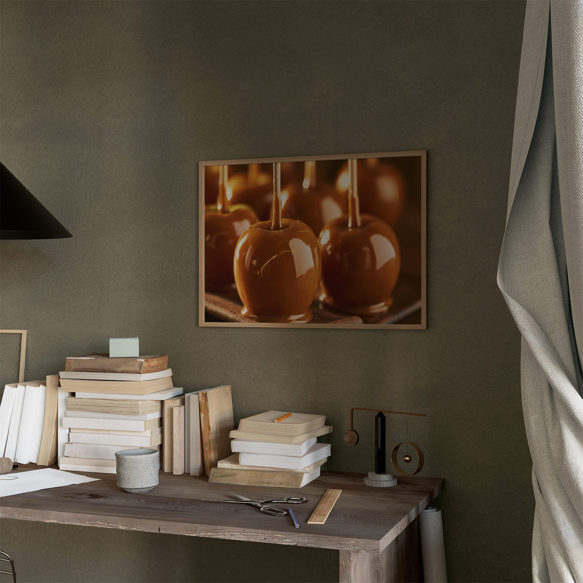 Caramel Glow at Dusk, three glossy caramel glazed apples on sticks in warm candlelight, painted in amber, burnt sugar and deep brown, in a thin wood frame on a moss green wall above a desk stacked with books