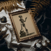 Class Is Still in Session, vintage engraving style skeleton lecturing atop a stack of books and a skull on sepia paper, in an antique gold frame laid on white linen with ferns and a pocket watch