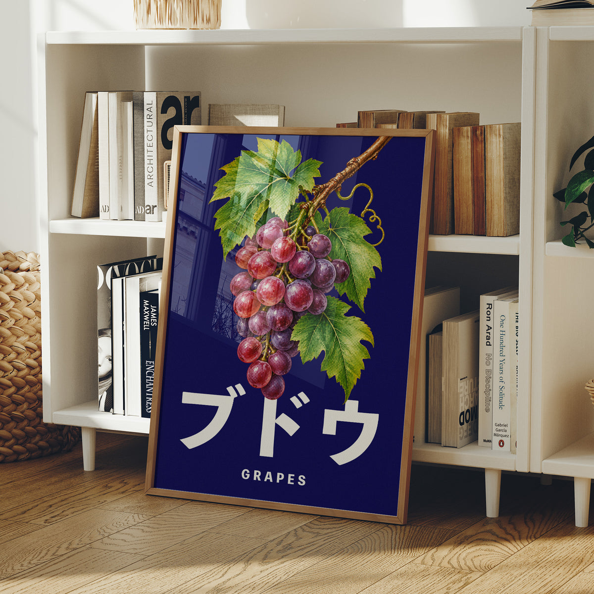 Clusters Heavy Over Midnight Water, a cluster of rose and purple grapes with green leaves hanging from a stem on a deep navy ground, with the Japanese katakana set in large white letters and the word GRAPES in small capitals beneath, in a wood frame on a white bookshelf beside stacked books and a small basket