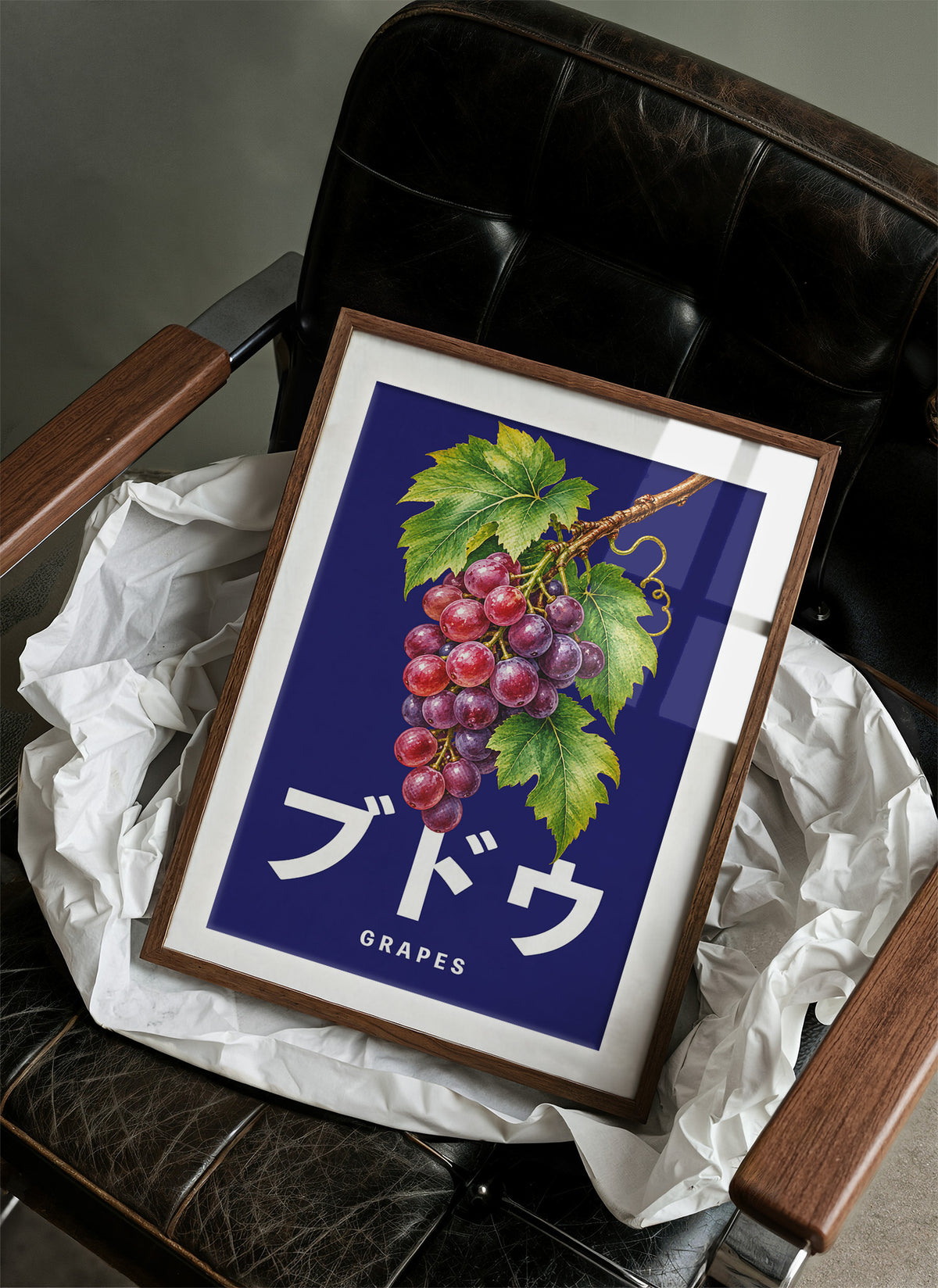 Clusters Heavy Over Midnight Water, a cluster of rose and purple grapes with green leaves hanging from a stem on a deep navy ground, with the Japanese katakana set in large white letters and the word GRAPES in small capitals beneath, in a wood frame on a black leather armchair beside crumpled white tissue