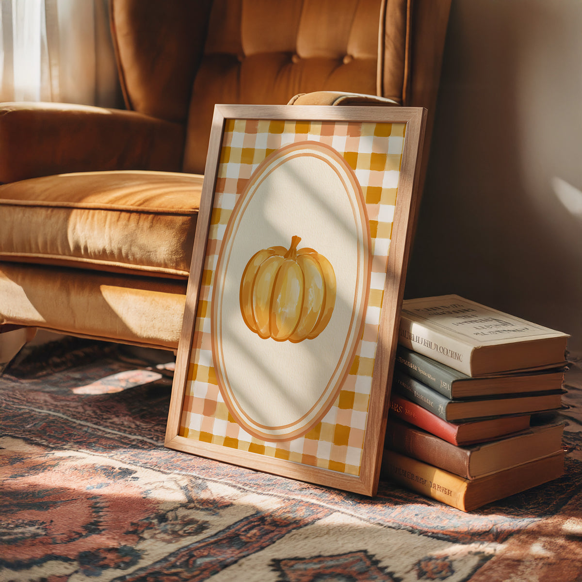 Dressed Up for a Quiet Afternoon, a single orange pumpkin in a cream oval medallion framed by a yellow and rust gingham border, painted in mustard, cream and burnt orange, in a wood frame on a rug beside a leather armchair and a stack of books