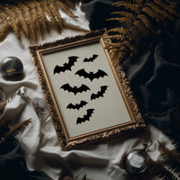 Evening Flight in Silhouette, a loose flock of black bat silhouettes spread across a cream ground in a simple, flat graphic arrangement, in an ornate frame on silk cloth beside ferns and a brass compass