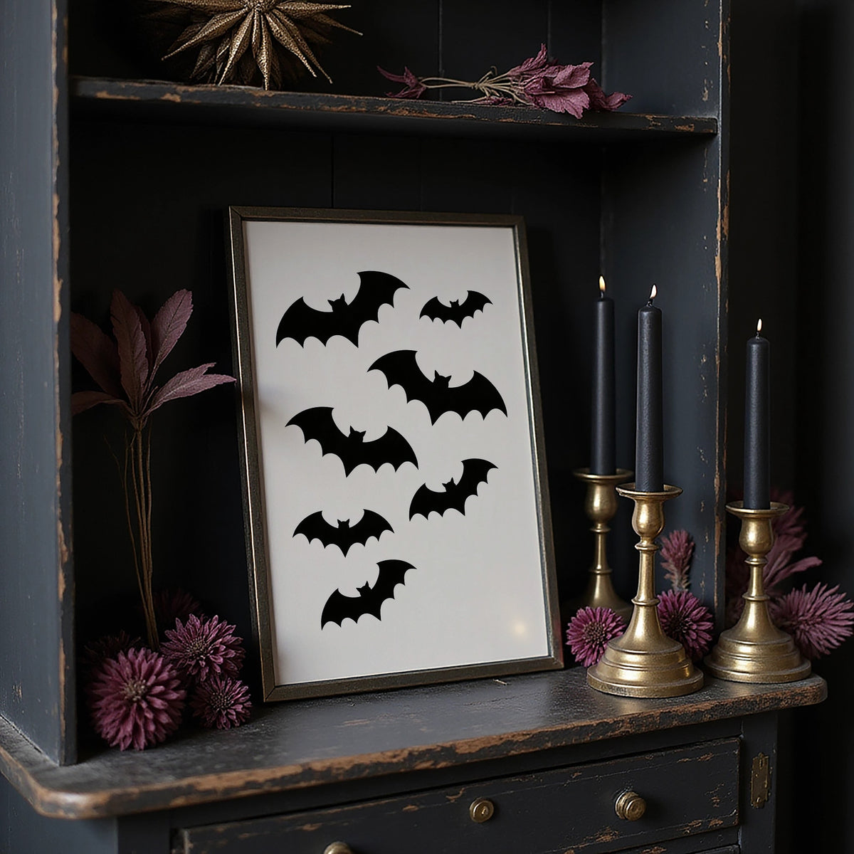 Evening Flight in Silhouette, a loose flock of black bat silhouettes spread across a cream ground in a simple, flat graphic arrangement, in a dark frame on a black shelf beside brass candles and dried leaves