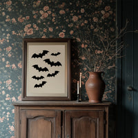 Evening Flight in Silhouette, a loose flock of black bat silhouettes spread across a cream ground in a simple, flat graphic arrangement, in a dark wood frame on a floral wallpaper wall above a rustic cabinet and a vase