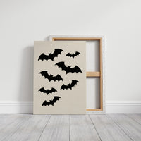 Evening Flight in Silhouette, a loose flock of black bat silhouettes spread across a cream ground in a simple, flat graphic arrangement, on a gallery canvas with wrapped edges leaning against a white wall