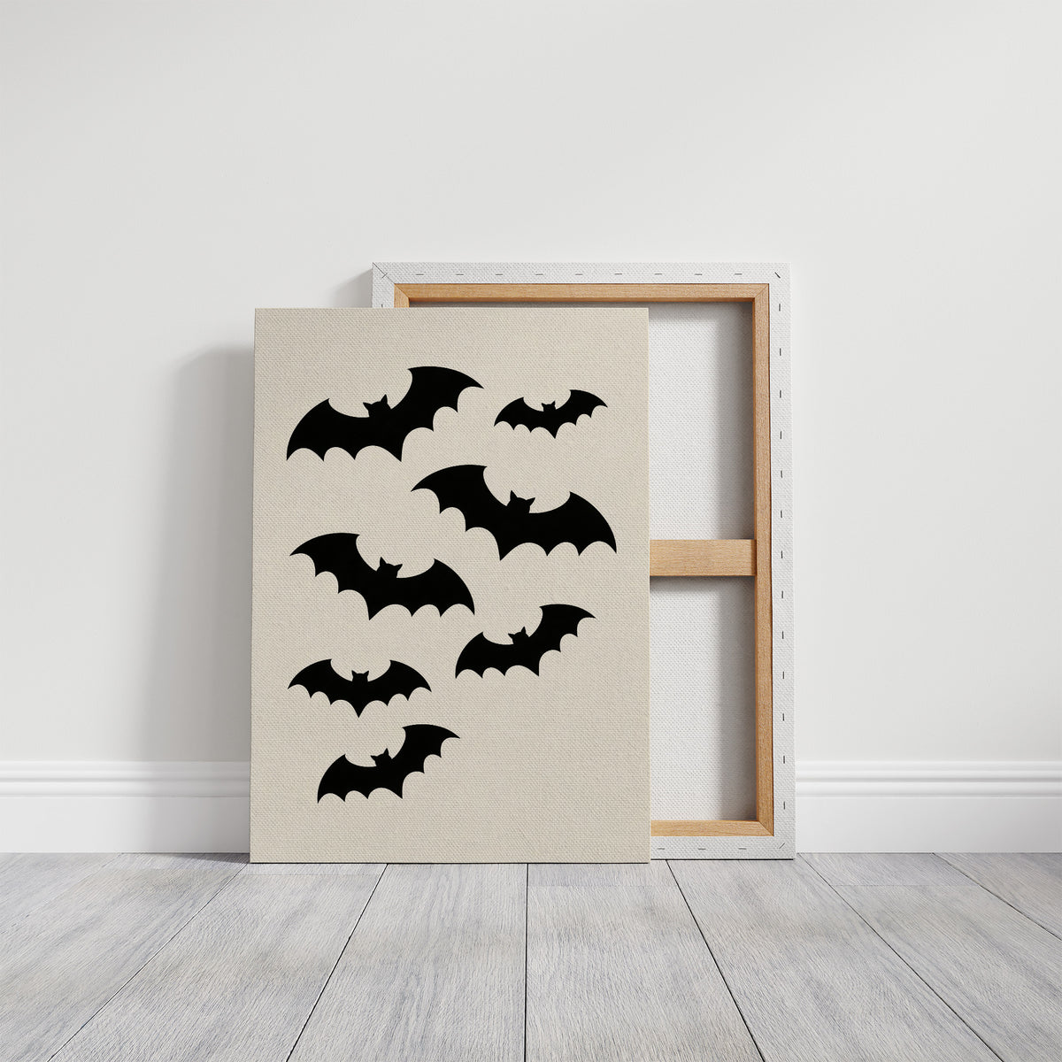 Evening Flight in Silhouette, a loose flock of black bat silhouettes spread across a cream ground in a simple, flat graphic arrangement, on a gallery canvas with wrapped edges leaning against a white wall