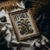 Every Head Grows Back Bolder, a many headed serpent with blue, red and gold scales rising in fans of heads, in a bold flash sheet style with a black lettered name across the top on a cream ground, in an ornate frame on silk cloth beside ferns and a brass compass