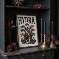 Every Head Grows Back Bolder, a many headed serpent with blue, red and gold scales rising in fans of heads, in a bold flash sheet style with a black lettered name across the top on a cream ground, in a dark frame on a black shelf beside brass candles and dried leaves