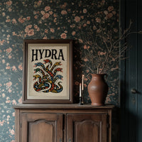Every Head Grows Back Bolder, a many headed serpent with blue, red and gold scales rising in fans of heads, in a bold flash sheet style with a black lettered name across the top on a cream ground, in a dark wood frame on a floral wallpaper wall above a rustic cabinet and a vase