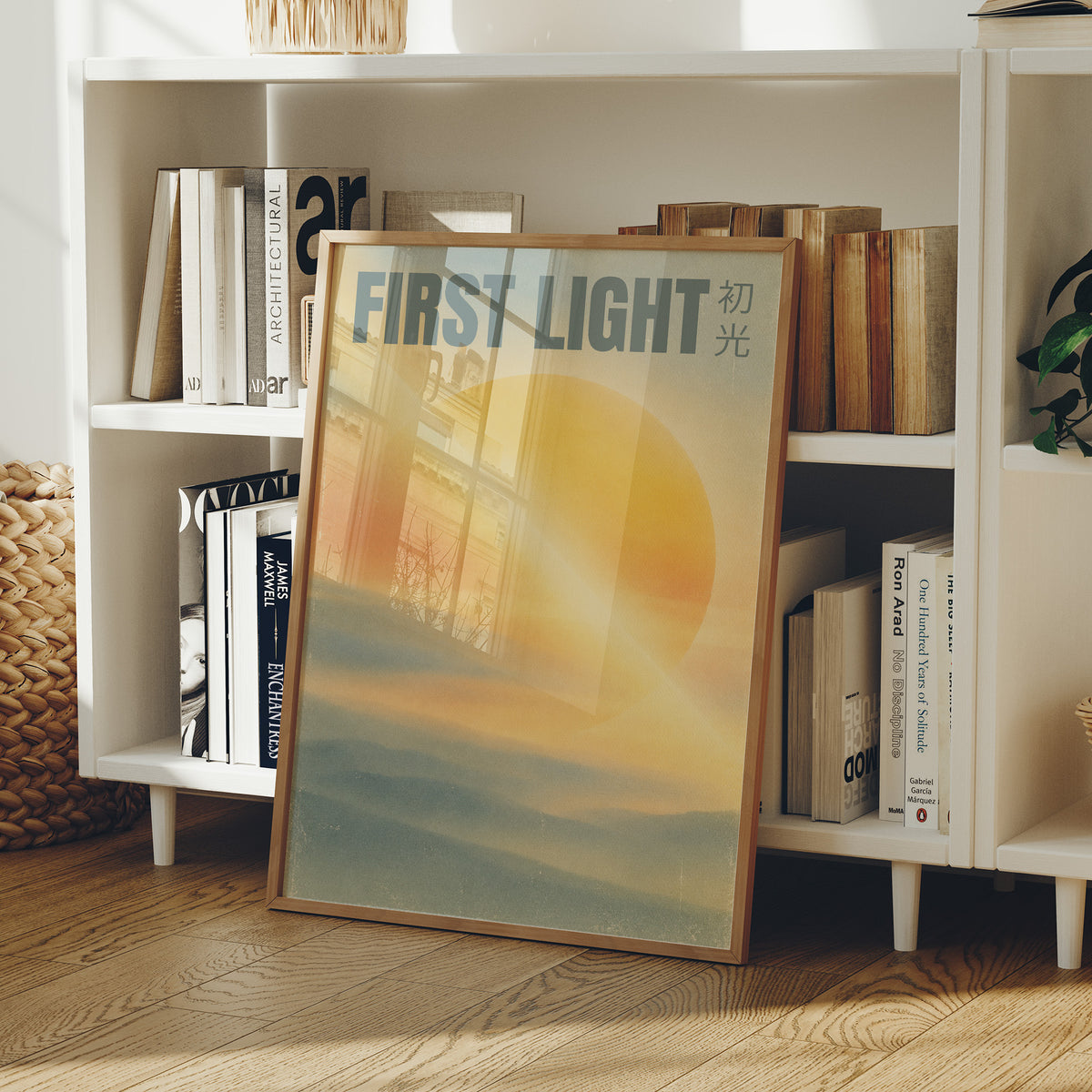 First Light Over a Quiet Sea, a pale sunrise over a soft sea, a warm yellow sun glowing above a band of blue water and peach clouds, with the words FIRST LIGHT in gray capitals across the top and a small Japanese word beside them, in a wood frame on a white bookshelf beside stacked books and a small basket