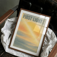 First Light Over a Quiet Sea, a pale sunrise over a soft sea, a warm yellow sun glowing above a band of blue water and peach clouds, with the words FIRST LIGHT in gray capitals across the top and a small Japanese word beside them, in a wood frame on a black leather armchair beside crumpled white tissue