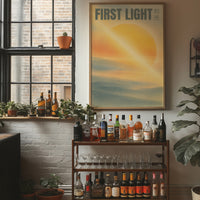 First Light Over a Quiet Sea, a pale sunrise over a soft sea, a warm yellow sun glowing above a band of blue water and peach clouds, with the words FIRST LIGHT in gray capitals across the top and a small Japanese word beside them, in a wood frame on a brick wall above a bar cart of bottles and plants