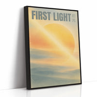First Light Over a Quiet Sea, a pale sunrise over a soft sea, a warm yellow sun glowing above a band of blue water and peach clouds, with the words FIRST LIGHT in gray capitals across the top and a small Japanese word beside them, as a framed canvas in a black frame