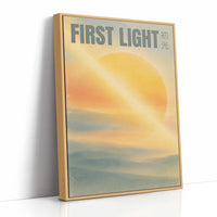 First Light Over a Quiet Sea, a pale sunrise over a soft sea, a warm yellow sun glowing above a band of blue water and peach clouds, with the words FIRST LIGHT in gray capitals across the top and a small Japanese word beside them, as a framed canvas in a natural wood frame