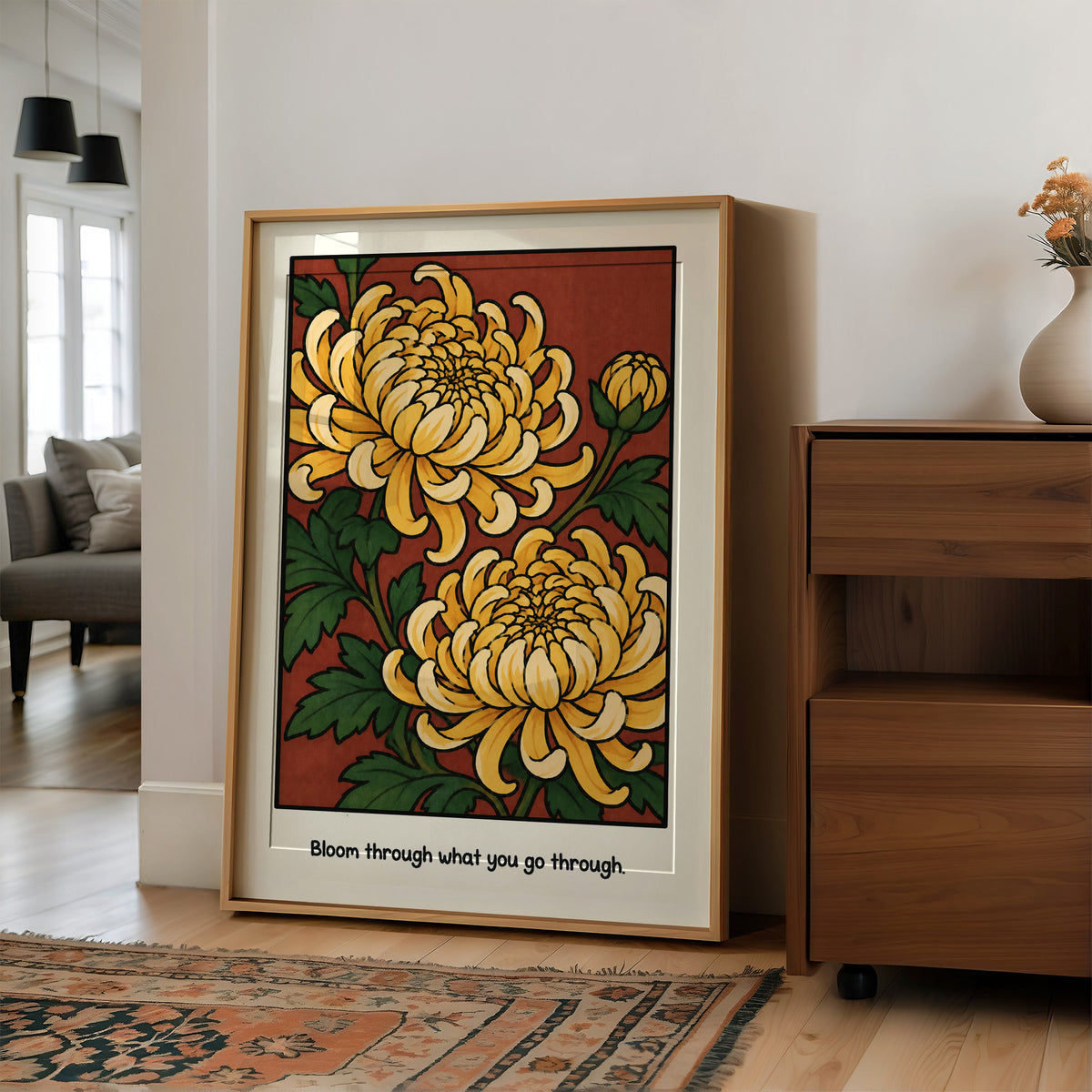 Gold Chrysanthemums Holding Their Ground, a cluster of yellow chrysanthemum blooms with green leaves and a bud on a rust red ground, with the words Bloom through what you go through in small black lettering along the bottom, in a wood frame on a wall beside a walnut cabinet and an open doorway