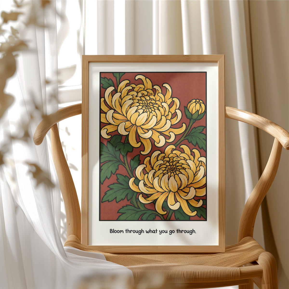 Gold Chrysanthemums Holding Their Ground, a cluster of yellow chrysanthemum blooms with green leaves and a bud on a rust red ground, with the words Bloom through what you go through in small black lettering along the bottom, in a wood frame beside a wicker chair by a sunny window