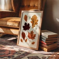 Gold Keeps Its Shape, a cluster of pressed autumn leaves in rust, maroon and gold on a cream ground, drawn in a botanical journal style, in a wood frame on a rug beside a leather armchair and a stack of books
