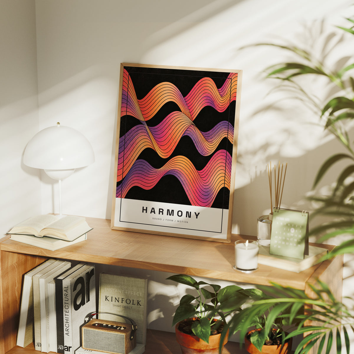 Harmony in Ember and Plum, rolling wavy bands of orange, magenta, violet and gold in fine contour lines on a deep black ground, with the word HARMONY in spaced black capitals on a cream label at the base, in a wood frame on a console table beside a white lamp and stacked books