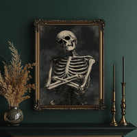 He Keeps His Arms Folded, a seated skeleton with crossed arms and bare ribs against a smoky charcoal ground, painted in bone, charcoal and ash gray, in an ornate gold frame on a deep green wall beside tall brass candlesticks