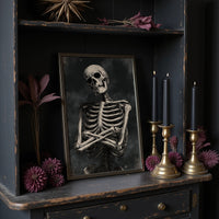 He Keeps His Arms Folded, a seated skeleton with crossed arms and bare ribs against a smoky charcoal ground, painted in bone, charcoal and ash gray, in a dark frame on a black shelf beside brass candles and dried leaves
