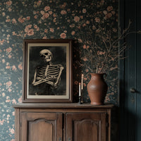 He Keeps His Arms Folded, a seated skeleton with crossed arms and bare ribs against a smoky charcoal ground, painted in bone, charcoal and ash gray, in a dark wood frame on a floral wallpaper wall above a rustic cabinet and a vase
