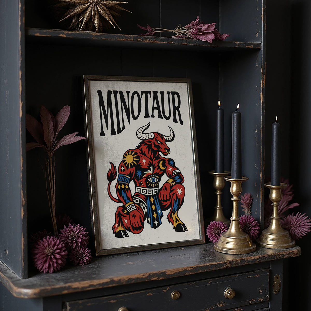 Horns in the Labyrinth, a red and patterned minotaur seated with a braced stance and a horned head, painted in bold black outlines on a cream paper ground beneath bold black lettering, in a traditional tattoo flash style, in a dark frame on a black shelf beside brass candles and dried leaves