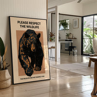 Keep a Respectful Distance, a large black bear with amber and brown fur, head low and shoulders bunched, set on a tan ground under bold black lettering that reads PLEASE RESPECT THE WILDLIFE in a vintage warning sign style, in a black frame leaning on a bright hallway floor beside a potted plant