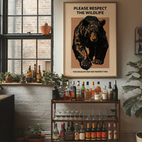 Keep a Respectful Distance, a large black bear with amber and brown fur, head low and shoulders bunched, set on a tan ground under bold black lettering that reads PLEASE RESPECT THE WILDLIFE in a vintage warning sign style, in a wood frame on a brick wall above a bar cart of bottles and plants