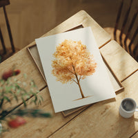 Keeping Its Warmth a Little Longer, a single watercolor tree in amber and rust with a soft brown trunk on a cream ground, the leaves painted in loose soft strokes, lying flat on a light wood table beside a coffee cup and red berries