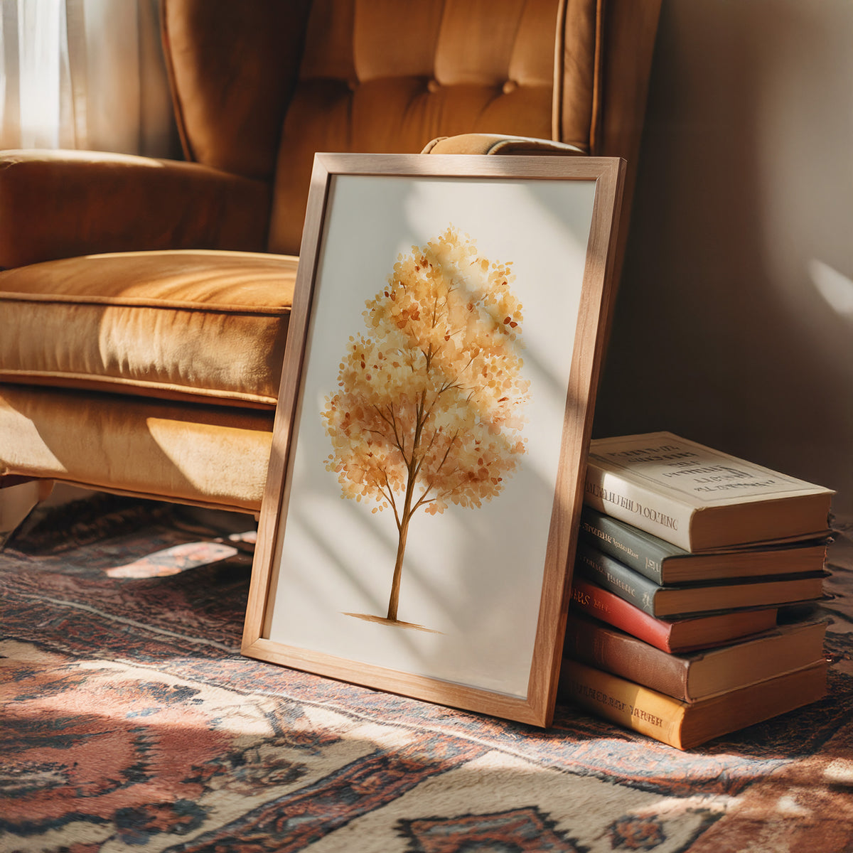Keeping Its Warmth a Little Longer, a single watercolor tree in amber and rust with a soft brown trunk on a cream ground, the leaves painted in loose soft strokes, in a wood frame on a rug beside a leather armchair and a stack of books