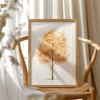 Keeping Its Warmth a Little Longer, a single watercolor tree in amber and rust with a soft brown trunk on a cream ground, the leaves painted in loose soft strokes, in a wood frame beside a wicker chair by a sunny window