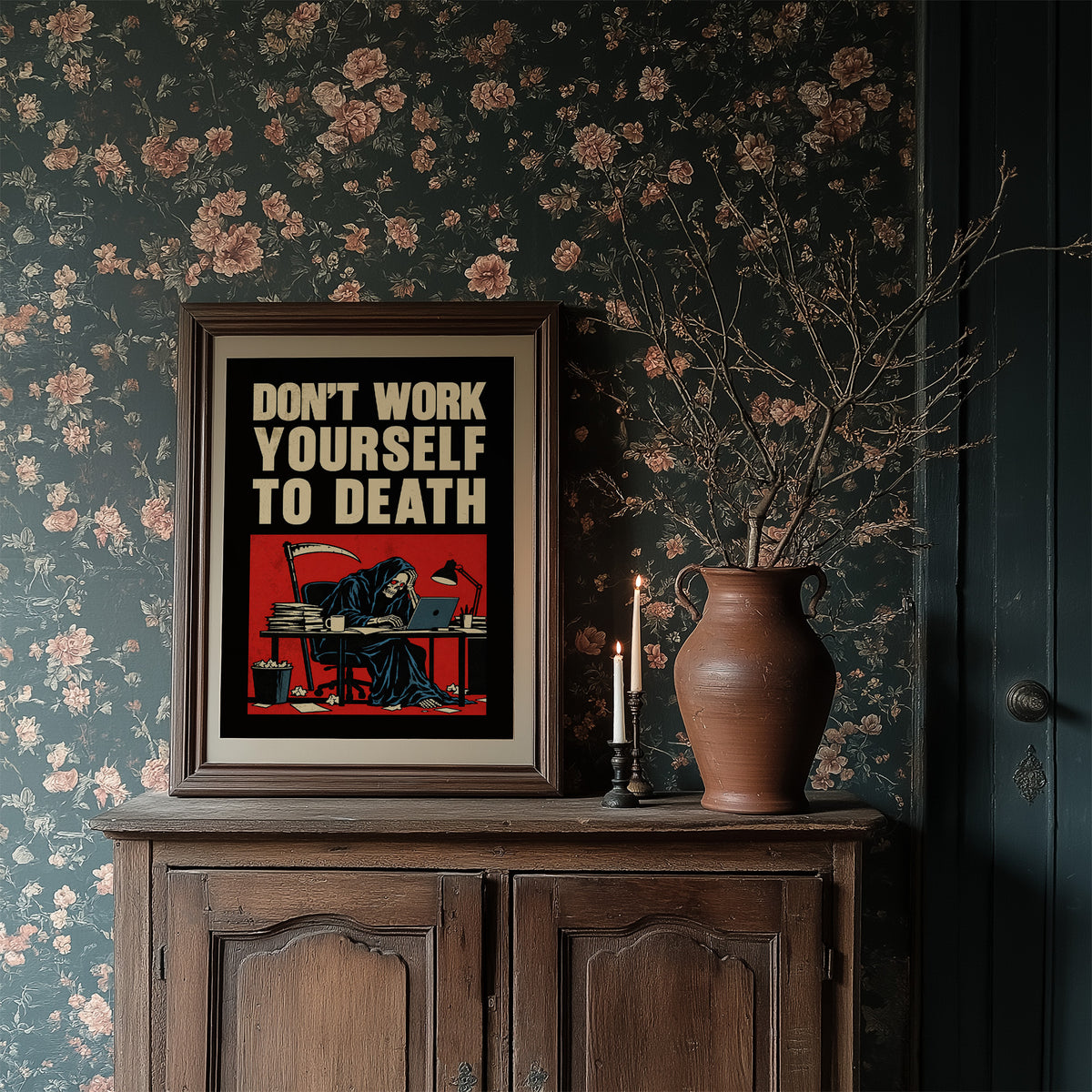 Let the Inbox Wait Till Monday, a hooded reaper hunched over a cluttered desk with a laptop and a stack of files, in a red panel with the lettering DON'T WORK YOURSELF TO DEATH in heavy cream capitals on black, in a dark wood frame on a floral wallpaper wall above a rustic cabinet and a vase