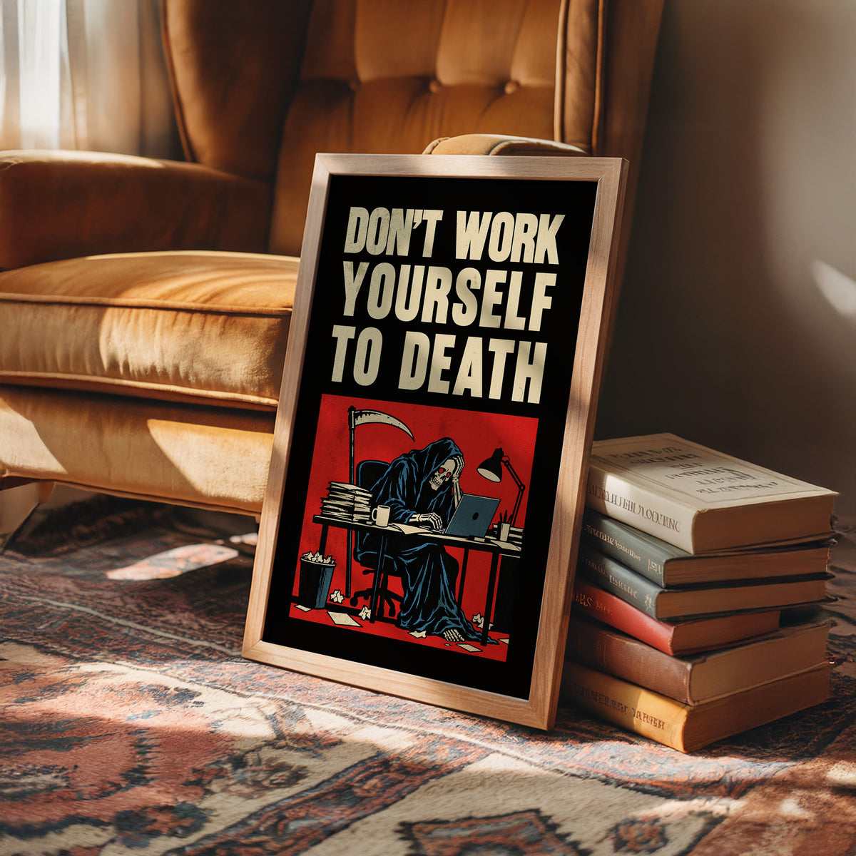 Let the Inbox Wait Till Monday, a hooded reaper hunched over a cluttered desk with a laptop and a stack of files, in a red panel with the lettering DON'T WORK YOURSELF TO DEATH in heavy cream capitals on black, in a wood frame on a leather armchair beside a stack of books