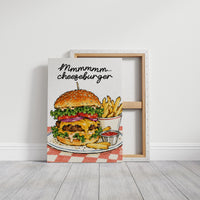 Mmmm Cheeseburger Makes Everything Better, a stacked cheeseburger with lettuce, tomato, cheese and a sesame bun beside fries and ketchup cups on a red and white checkered plate, with the lettering Mmmmm cheeseburger in black script across the top, on a gallery canvas with wrapped edges leaning against a white wall