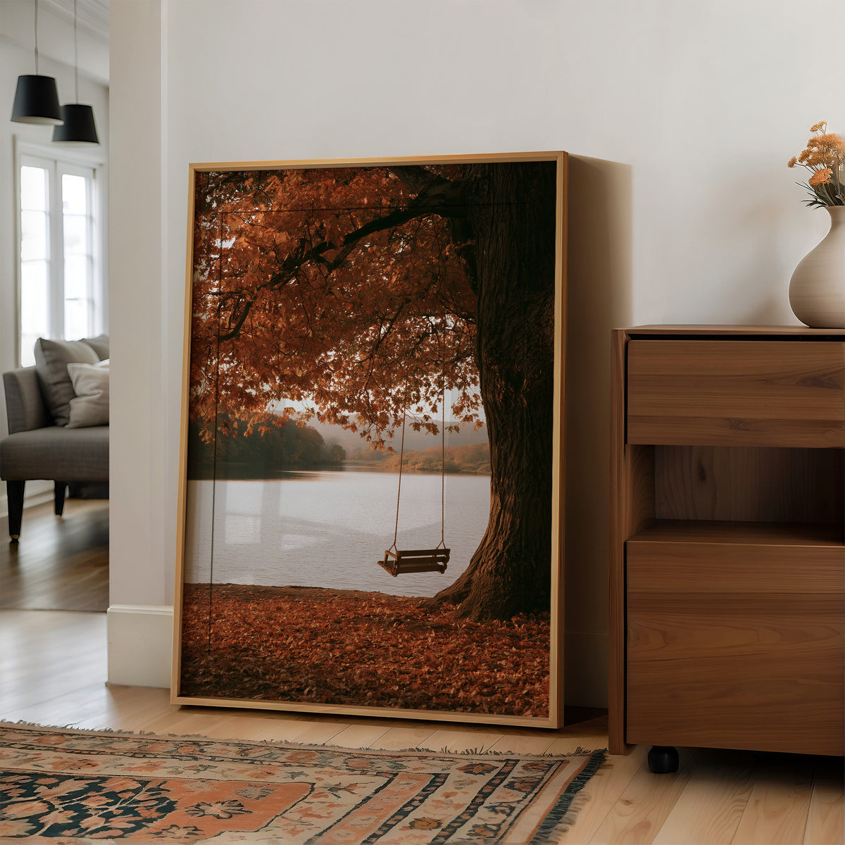 Nobody Has Swung Since Morning, a lone wooden swing hanging from a great tree with orange leaves above a still lake, painted in burnt orange, rust and misty gray, in a wood frame on a wall beside a walnut cabinet and an open doorway