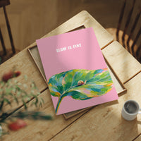 Nothing Here Is Rushed, a green leaf curling across the lower half of a blush pink ground with a small snail resting on its curve, with a pale line of lettering above, in a loose watercolor style painted in blush pink, green and soft gold, lying flat on a light wood table beside a coffee cup and red berries