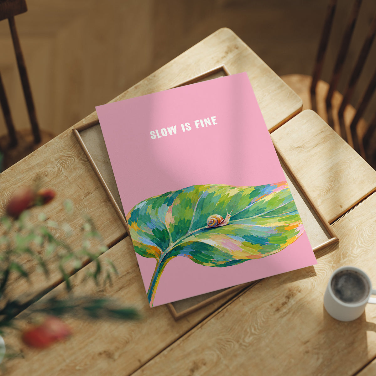Nothing Here Is Rushed, a green leaf curling across the lower half of a blush pink ground with a small snail resting on its curve, with a pale line of lettering above, in a loose watercolor style painted in blush pink, green and soft gold, lying flat on a light wood table beside a coffee cup and red berries