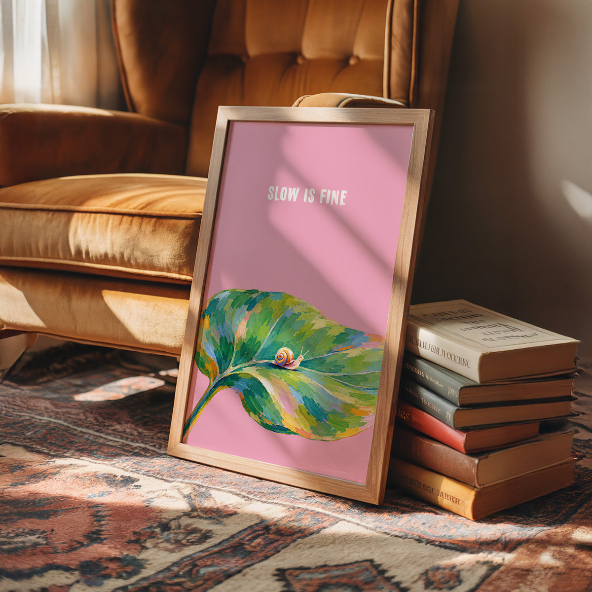 Nothing Here Is Rushed, a green leaf curling across the lower half of a blush pink ground with a small snail resting on its curve, with a pale line of lettering above, in a loose watercolor style painted in blush pink, green and soft gold, in a wood frame on a rug beside a leather armchair and a stack of books