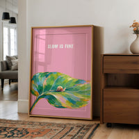 Nothing Here Is Rushed, a green leaf curling across the lower half of a blush pink ground with a small snail resting on its curve, with a pale line of lettering above, in a loose watercolor style painted in blush pink, green and soft gold, in a wood frame on a wall beside a walnut cabinet and an open doorway
