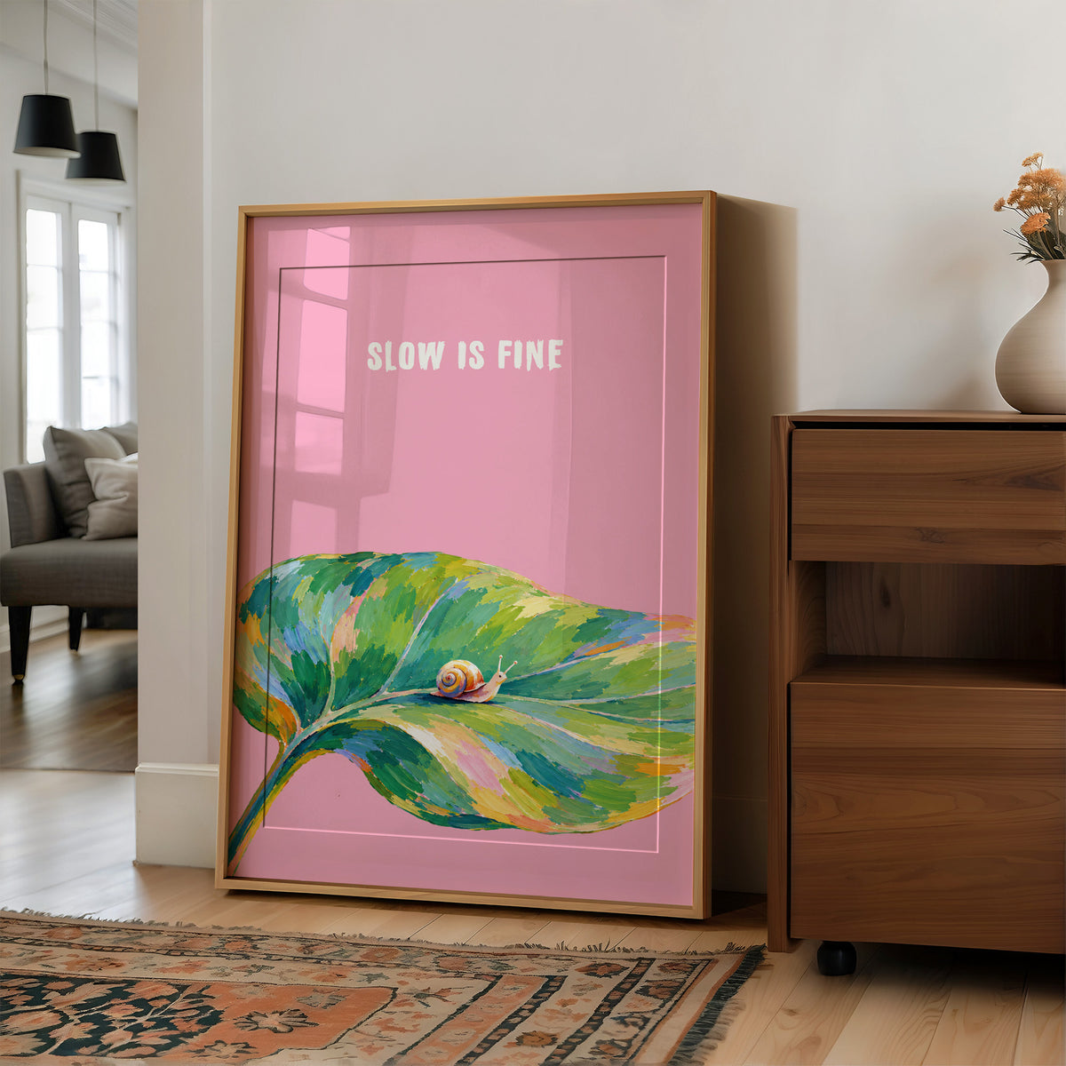 Nothing Here Is Rushed, a green leaf curling across the lower half of a blush pink ground with a small snail resting on its curve, with a pale line of lettering above, in a loose watercolor style painted in blush pink, green and soft gold, in a wood frame on a wall beside a walnut cabinet and an open doorway