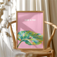 Nothing Here Is Rushed, a green leaf curling across the lower half of a blush pink ground with a small snail resting on its curve, with a pale line of lettering above, in a loose watercolor style painted in blush pink, green and soft gold, in a wood frame beside a wicker chair by a sunny window