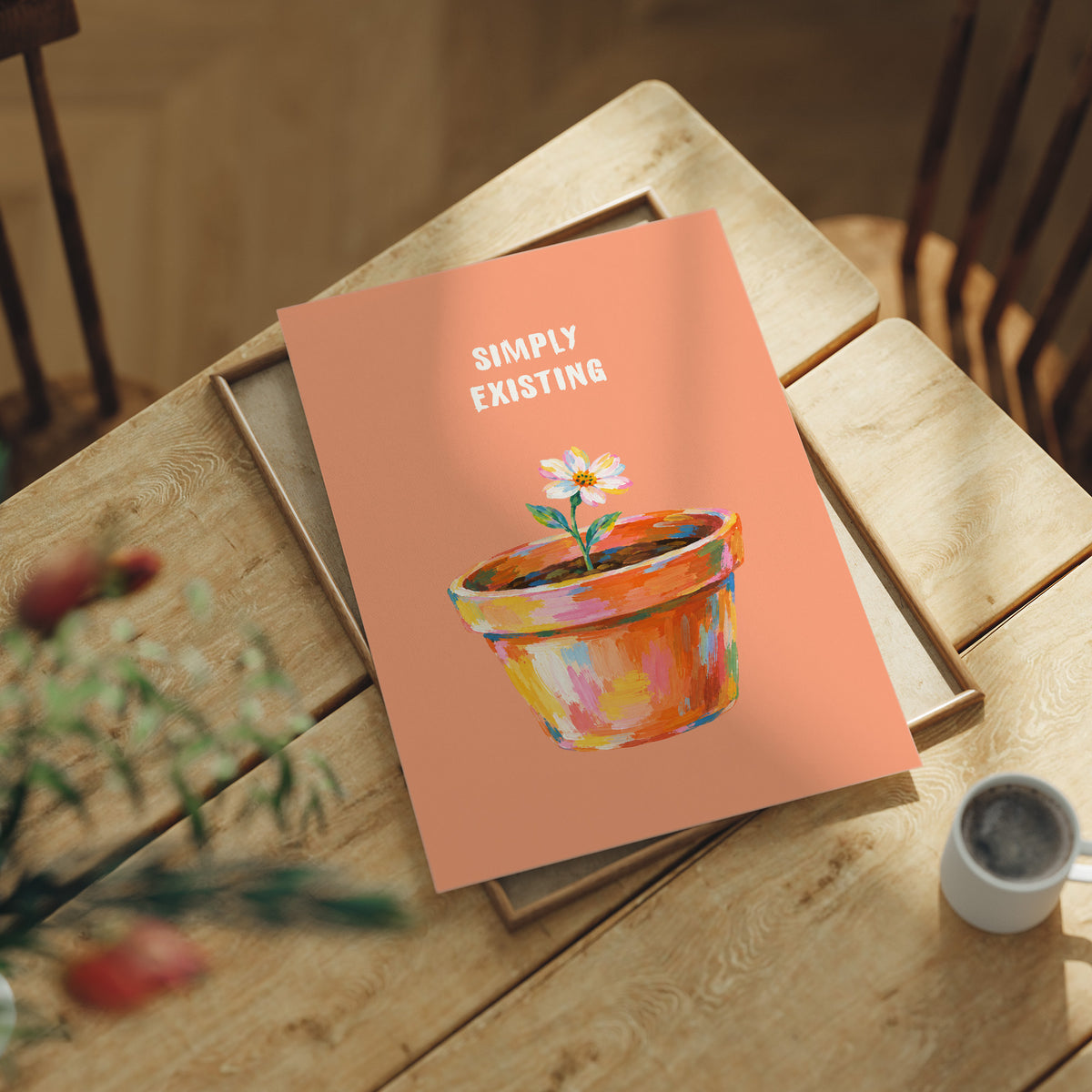 One Small Bloom in Clay, a single white flower rising from a painted clay pot in coral, gold and pink brushstrokes, on a peach ground with two lines of bold pale lettering above, in a loose painterly style, lying flat on a light wood table beside a coffee cup and red berries
