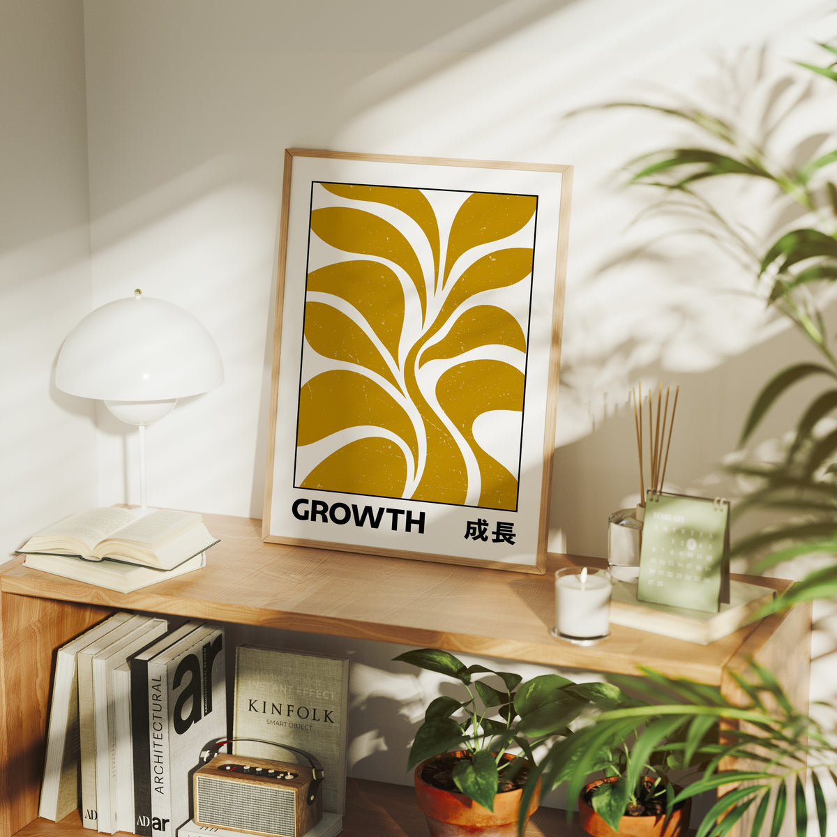 Patient Leaves Turning Toward Light, a stylized mustard leaf motif in curved shapes on a cream ground, with the word GROWTH in black capitals and a small Japanese word beside it, in a wood frame on a console table beside a white lamp and stacked books