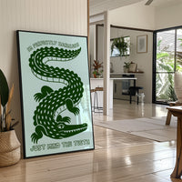 Perfectly Harmless, Mind the Teeth, a deep green crocodile curling down a pale mint ground, with a bold hand lettered line above reading I'M PERFECTLY HARMLESS and one below reading JUST MIND THE TEETH, in a black frame leaning on a bright hallway floor beside a potted plant