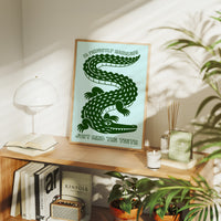 Perfectly Harmless, Mind the Teeth, a deep green crocodile curling down a pale mint ground, with a bold hand lettered line above reading I'M PERFECTLY HARMLESS and one below reading JUST MIND THE TEETH, in a wood frame on a console table beside a white lamp and stacked books