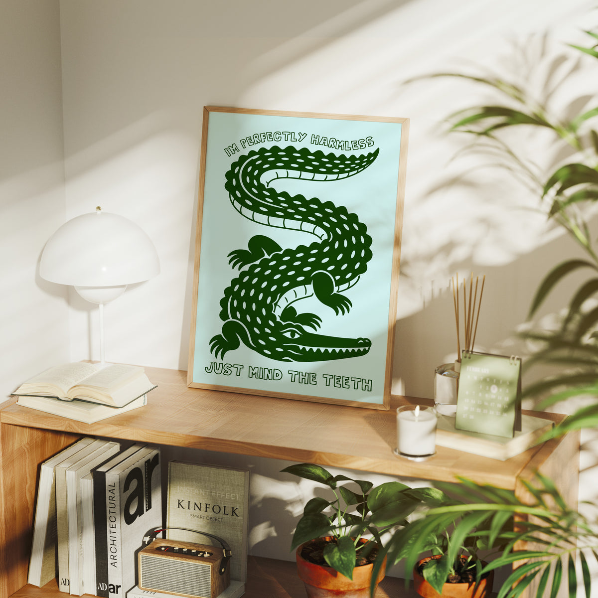 Perfectly Harmless, Mind the Teeth, a deep green crocodile curling down a pale mint ground, with a bold hand lettered line above reading I'M PERFECTLY HARMLESS and one below reading JUST MIND THE TEETH, in a wood frame on a console table beside a white lamp and stacked books