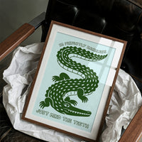 Perfectly Harmless, Mind the Teeth, a deep green crocodile curling down a pale mint ground, with a bold hand lettered line above reading I'M PERFECTLY HARMLESS and one below reading JUST MIND THE TEETH, in a wood frame on a black leather armchair beside crumpled white tissue