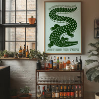 Perfectly Harmless, Mind the Teeth, a deep green crocodile curling down a pale mint ground, with a bold hand lettered line above reading I'M PERFECTLY HARMLESS and one below reading JUST MIND THE TEETH, in a wood frame on a brick wall above a bar cart of bottles and plants