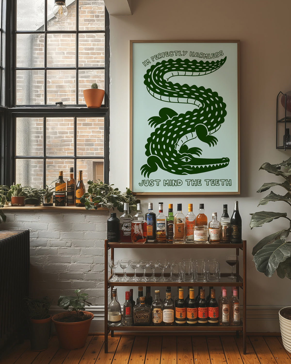 Perfectly Harmless, Mind the Teeth, a deep green crocodile curling down a pale mint ground, with a bold hand lettered line above reading I'M PERFECTLY HARMLESS and one below reading JUST MIND THE TEETH, in a wood frame on a brick wall above a bar cart of bottles and plants