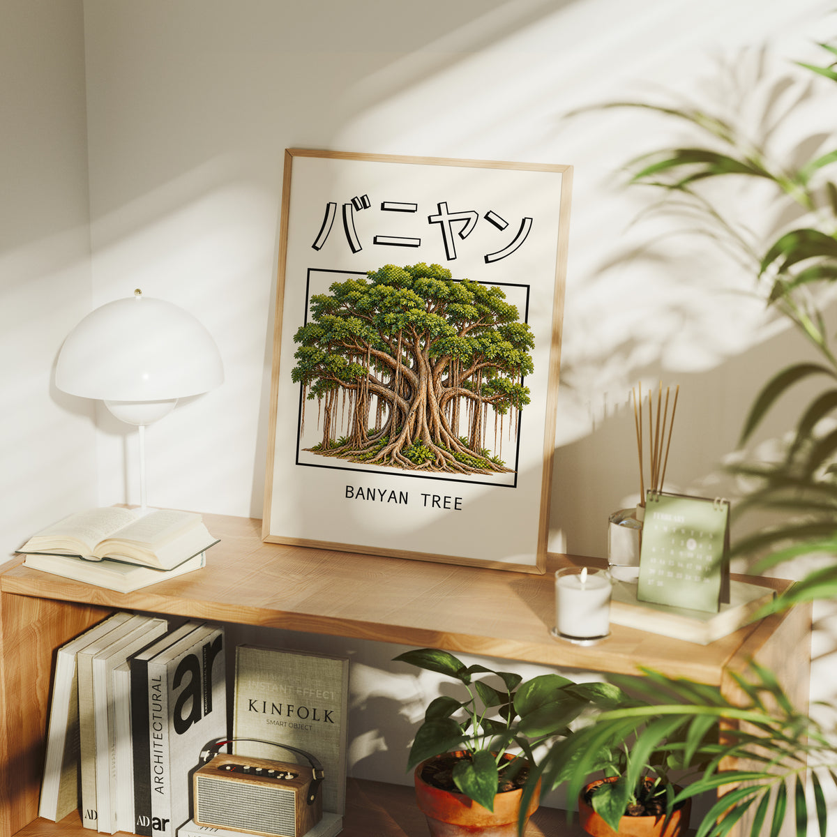 Roots That Remember the Ground, a banyan tree with a wide green canopy and many aerial roots hanging to the ground, drawn in a framed Japanese travel poster style with katakana above and BANYAN TREE below, in a wood frame on a console table beside a white lamp and stacked books